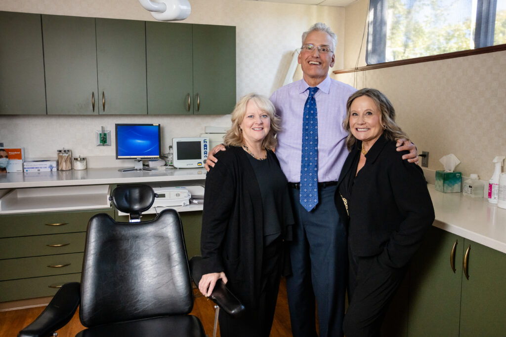 Three smiling doctors in professional attire are standing together in a cosmetic surgery medical office setting. 