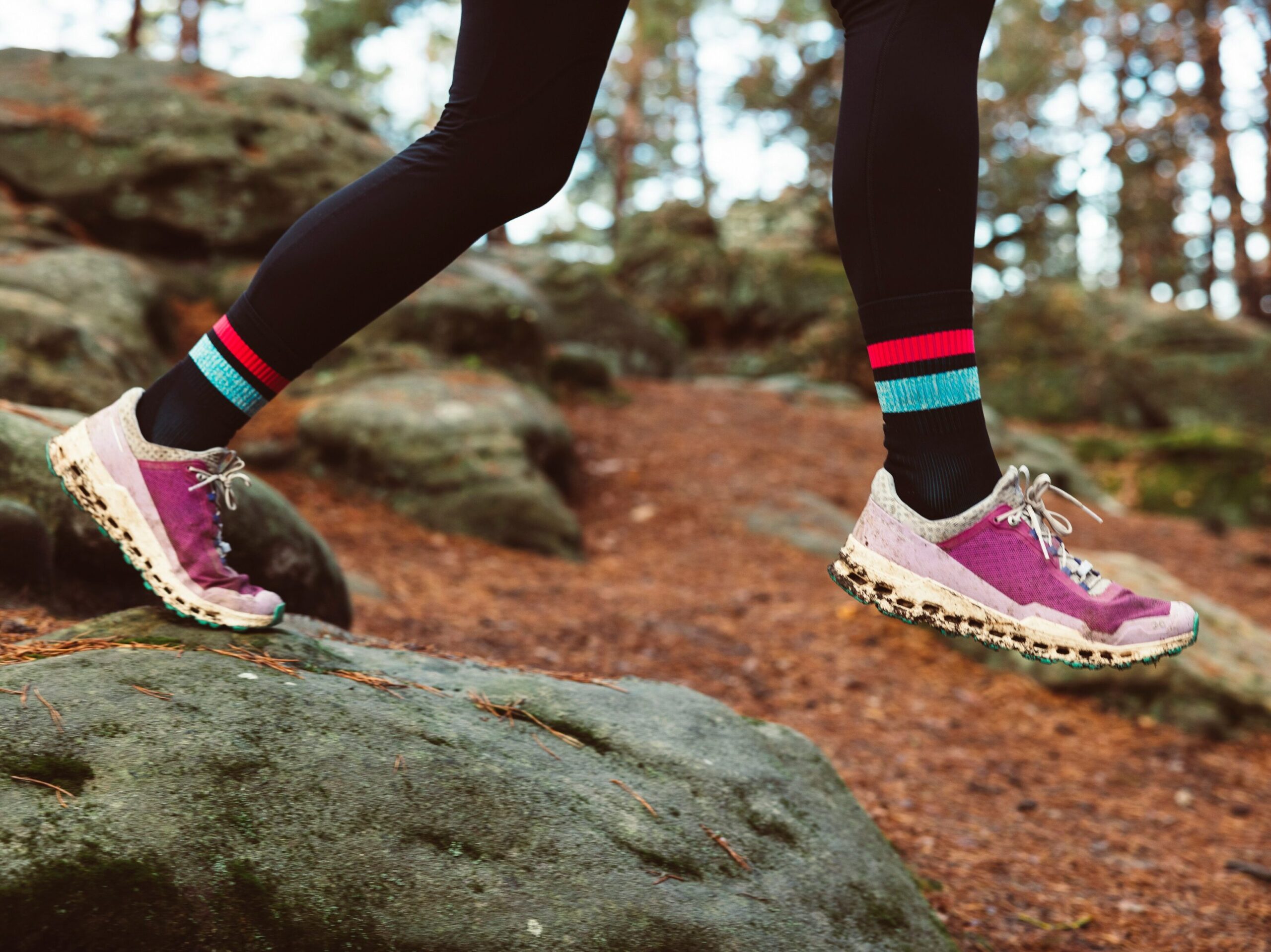 A runner, hopping over, rocks in the woods wearing black leggings and pink trail running shoes. 