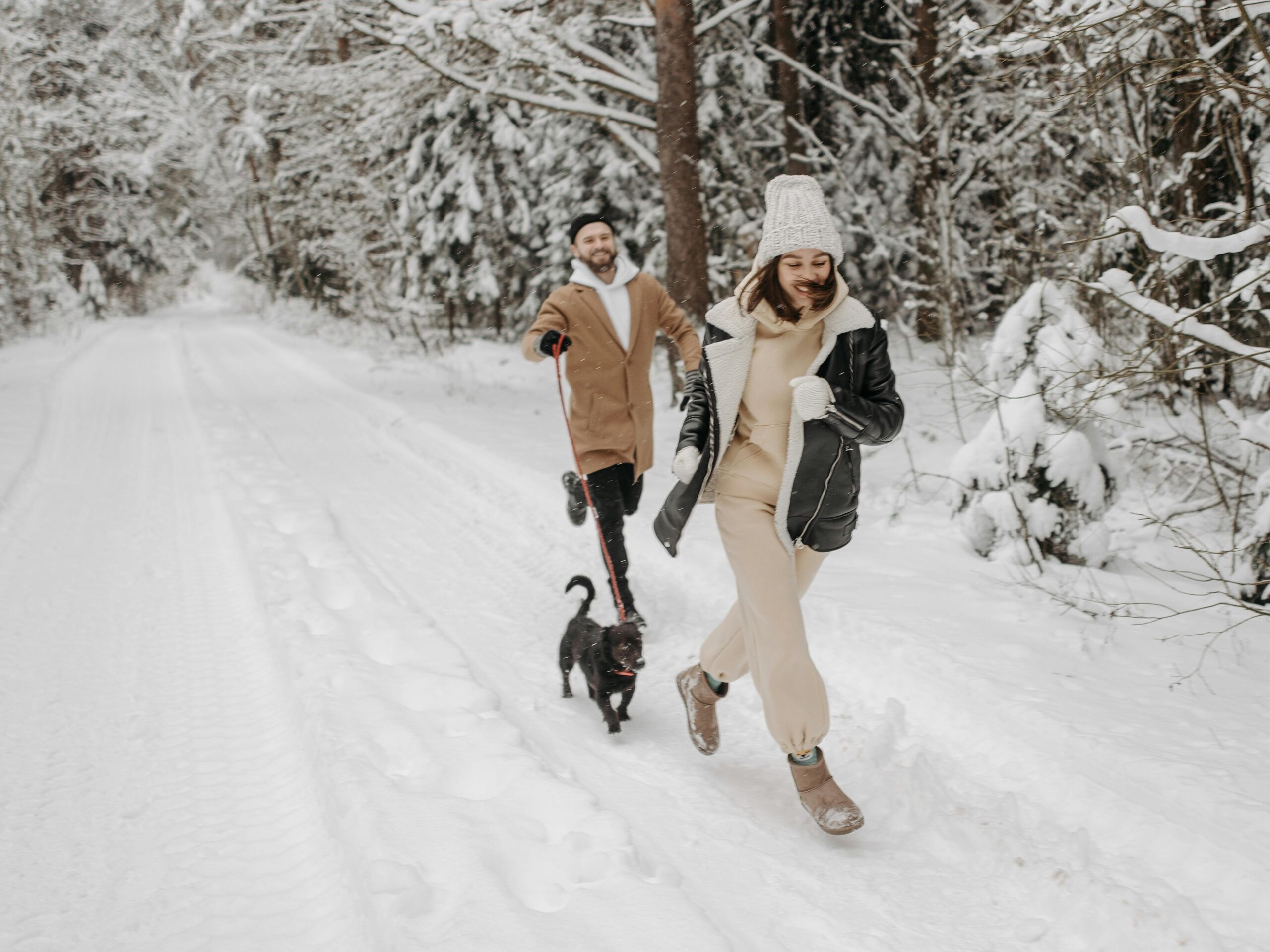 A man and woman happily trotting with their small dog through a snowy trail in the woods. 