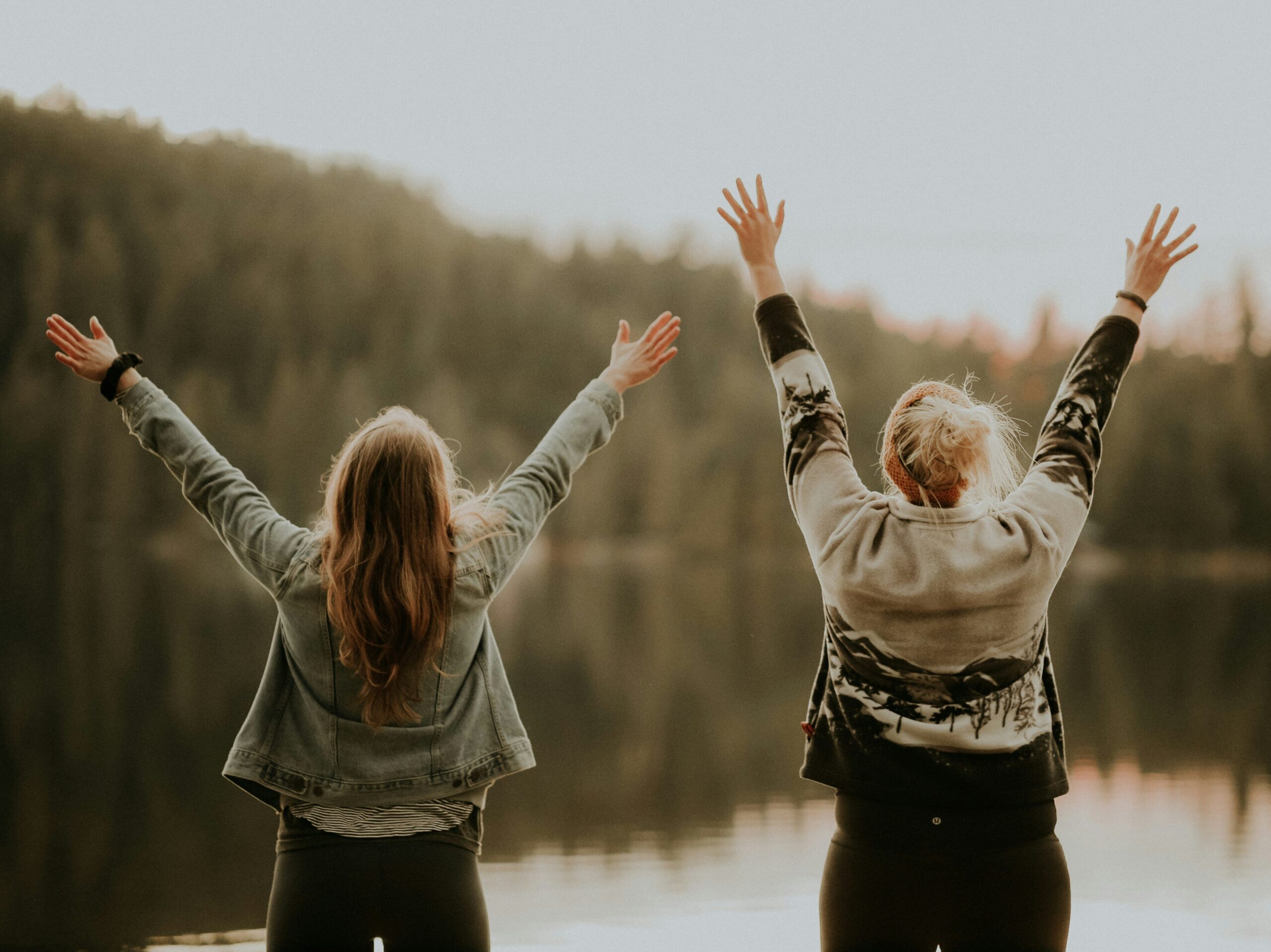 Two women are standing, facing a lake, with their hands in the air in celebration. 