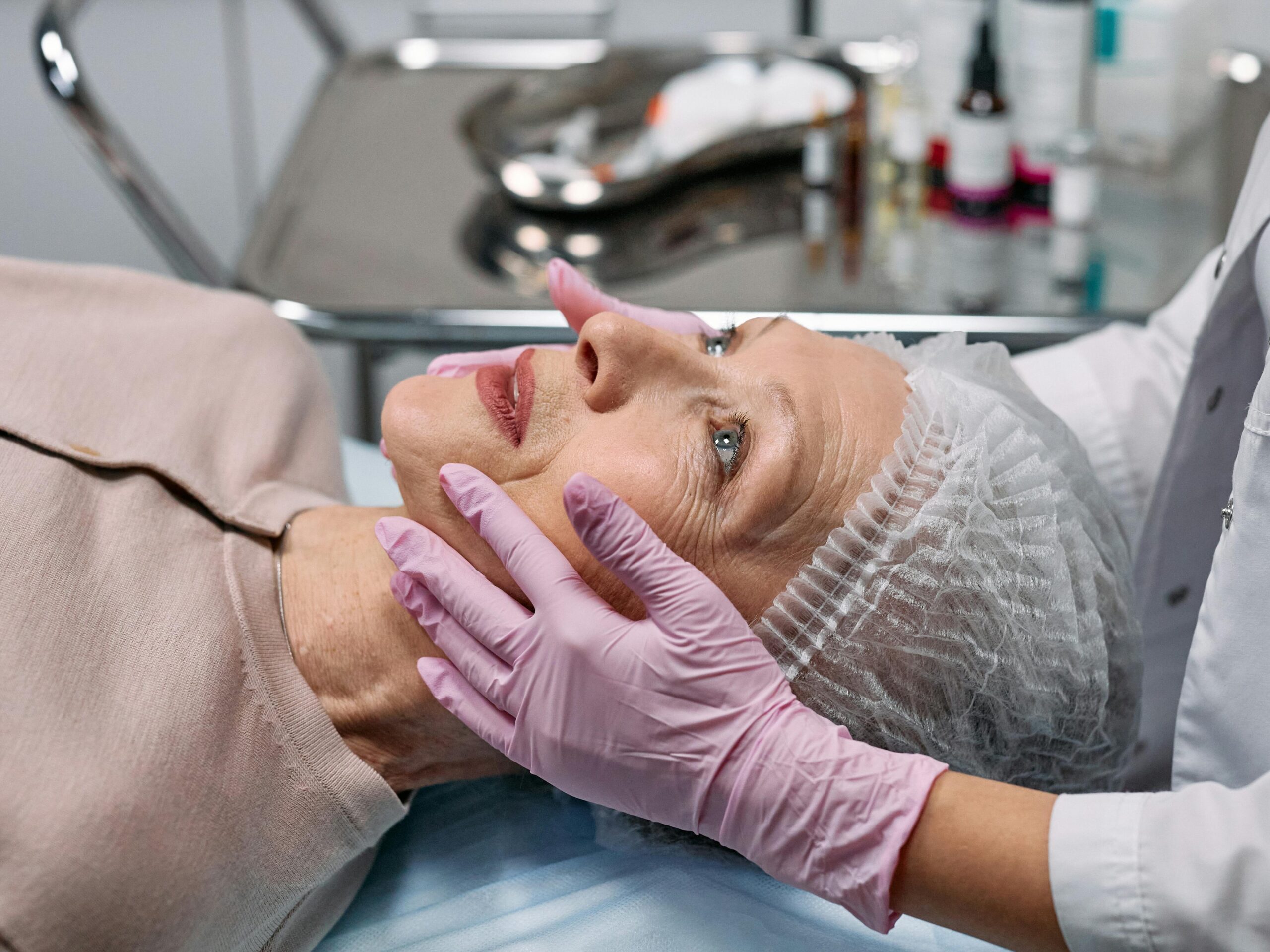 Woman lying in a hospital examination chair while a nurse examines her face, preparing for surgery.